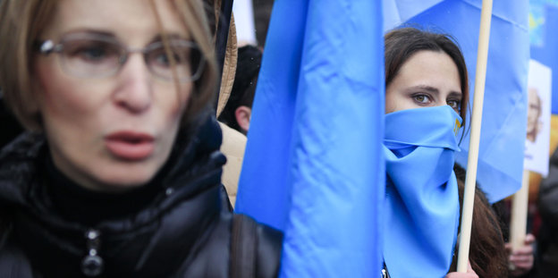 Zwei Frauen in warmen Winterjacken mit blauen, zusammengerollten Fahnen, eine hat auch eine blaue Fahne vor den Mund gebunden