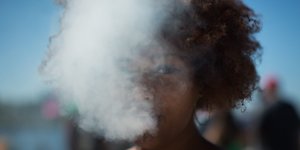 Eine Frau pustet dicken weißen Rauch aus, über ihr strahlt der blaue Himmel