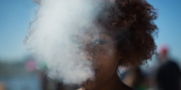 Eine Frau pustet dicken weißen Rauch aus, über ihr strahlt der blaue Himmel