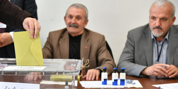 Eine Hand wirft einen gelben Umschlag in eine Plastikbox, im Hintergrund sehen zwei ältere Männer dabei zu