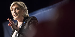 Marine Le Pen steht in Redner-Pose vor nachtschwarzem Hintergrund, nur ein Lichtstrahl fällt hinter ihrem Nacken durchs Bild