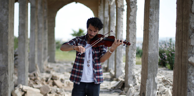 Ein junger Mann spielt auf seiner Geige. Er steht in einem Durchgang, im Hintergrund lassen sich Säulen und Schutt erkennen