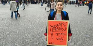 Eine Frau mit einem orangefarbenen Papschild steht auf einem Platz