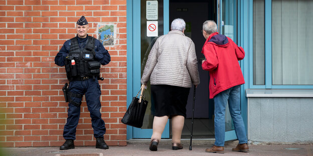 Eine ältere Frau mit Stock betritt durch eine Glastür ein Gebäude. Jemand öffnet ihr die Tür. Daneben steht ein Polizist, der das Gebäude bewacht