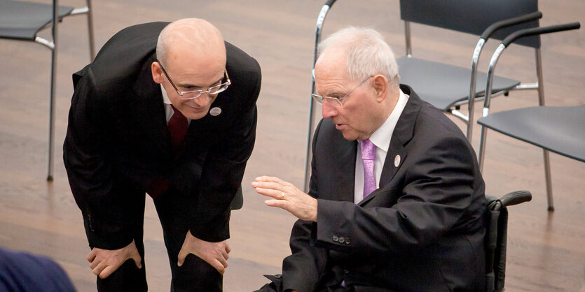 Zwei Männer in dunklen Anzügen unterhalten sich. Einer sitzt in einem Rollstuhl