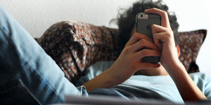 Ein Mann liegt im Bett und hält ein Handy in den Händen, sein Gesicht ist davon verdeckt