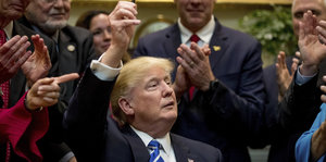 Donald Trump sitzt und grinst, um ihn herum stehen Menschen und applaudieren