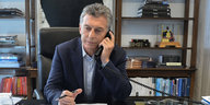 Macri sitzt an seinem Schreibtisch, mit dem Telefonhörer am Ohr, im Hintergrund ein BÜcherregal