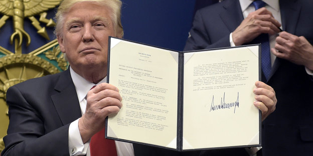 Trump hält Dokument mit seiner Unterschrift hoch und guckt siegessicher