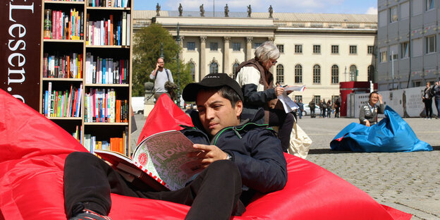 Ein junger Mann sitzt auf einem roten Sitzsack auf einem öffentlichen Platz und liest ein Magazin