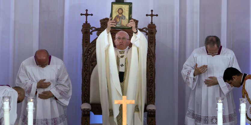 Ein Mann hält ein Bild hoch. Vor ihm steht ein Kreuz. Nehmen ihm verbeugen sich Männer.