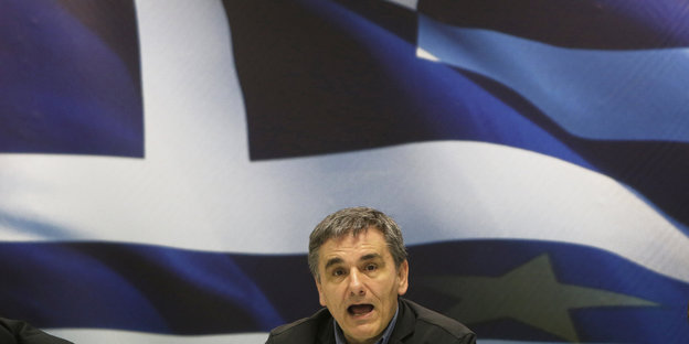 Der griechische Finanzminister Euklid Tsakalotos sitzt auf einer Pressekonferenz vor einer großen griechischen Flagge