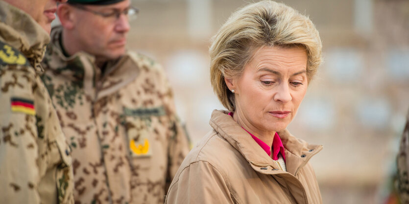 Ursula von der Leyen in sandfarbenem Anorak steht mit gesenktem Kopf vor zwei Soldaten