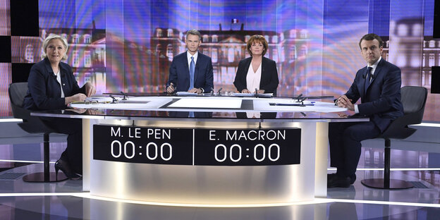 Marine Le Pen und Emmanuel MAcron sitzen an einem Tisch in einem TV-Studio. Zwischen ihnen sitzen eine Moderatorin und ein Moderator