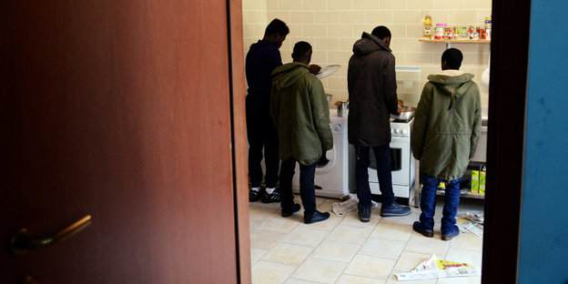 Vier junge Männer in zu großen Parkas stehen mit dem Rücken zum Betrachter verloren in einer Küche