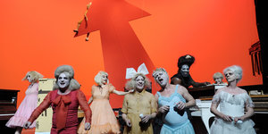 Im engen roten Kleid sieht man Ingo Günther zwischen anderen Schauspielern auf einer orangerot leuchtenden Bühne.