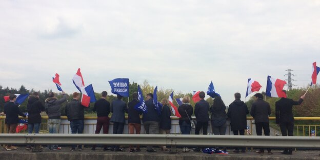 Mehrere junge Menschen stehen nebeneinander am Geländer einer Autobahnbrücke und wehen mit französischen Nationalflaggen