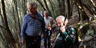 Drei ältere Männer stehen in einem schattigen Waldabschnitt, einer hält ein Gewehr