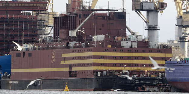 Ein großes Schiff in rotbraun mit beigen Streifen, dahinter Kräne