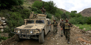 Ein Militär-Auto, daneben drei Soldaten, umgeben von grünen Wiesen und Büschen auf einem nicht geteerten Weg