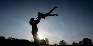 In der Abenddämmerung lässt ein Mann sein Kind, an beiden Händen fest gehalten, hoch in die Luft fliegen