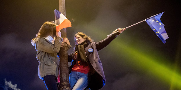 Zwei Personen halten sich an einem Laternenpfahl fest. Sie schwenken die französische und die Europaflagge