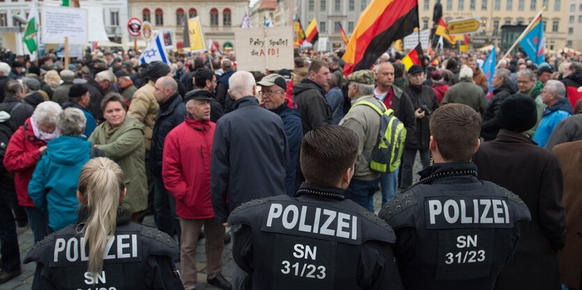 Im Vordergrund drei Polizisten, im Hintergrund ein Platz voller Menschen mit Plakaten und Flaggen