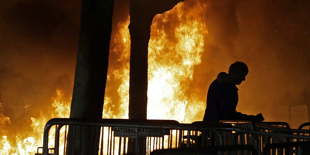 Hinter Absperrgittern Feuer lodert auf, ein Mensch steht daneben