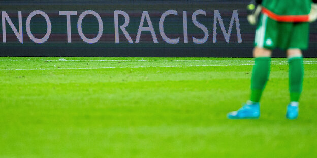 Ein Fußballspieler, dessen Oberkörper man nicht sieht, auf einem Fußballplatz vor einer Bande mit der Aufschrift "No to Racism".