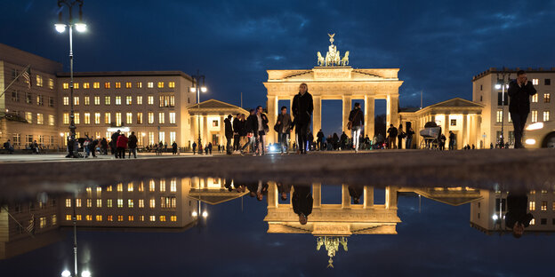 Menschen laufen im Dunkeln vor dem hell erleuteten Brandenburger Tor herum.