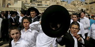 Menschen in orthodox-jüdischer Kleidung in Jerusalem. Im Vordergrund Kinder, ein Junge hält einen schwarzen Hut in die Kamera