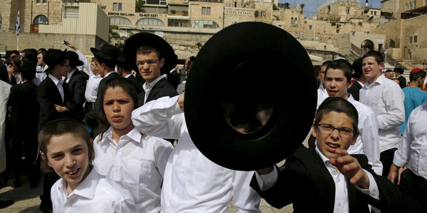 Menschen in orthodox-jüdischer Kleidung in Jerusalem. Im Vordergrund Kinder, ein Junge hält einen schwarzen Hut in die Kamera