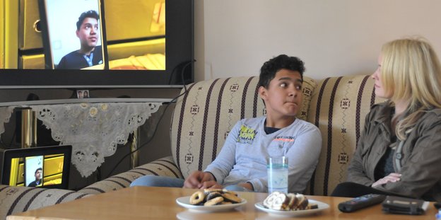 Zijush sitzt mit seiner Lehrerin in seinem Wohnzimmer in Skopje; im Hintergrund Fernseher mit Videoschalte in sein Bremerhavener Klassenzimmer