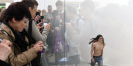 Besucher und Besucherinnen fotografieren eine Frau, die auf der Biennale in Venedig hinter Glas mit nacktem Oberkörper läuft