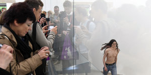 Besucher und Besucherinnen fotografieren eine Frau, die auf der Biennale in Venedig hinter Glas mit nacktem Oberkörper läuft