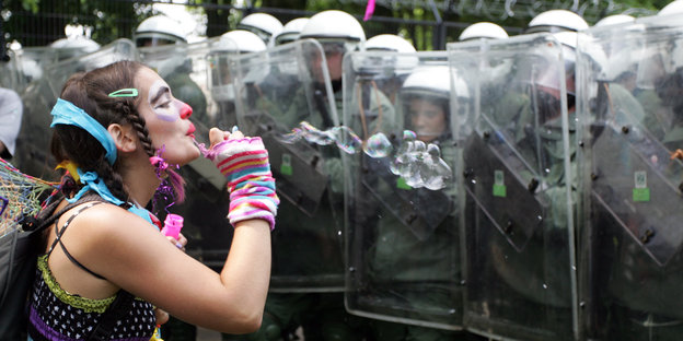 Eine als Clown geschminkte Frau bläst Seifenblasen gegen Plastikschilde, hinten denen Polizisten stehen.