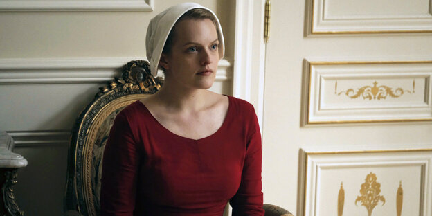 Offred (Elisabeth Moss) sitzt in einem roten Kleid und mit einem weißen Häubchen auf einem Sessel