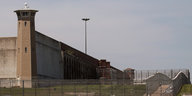 Hohe Gefängnismauern mit Wachturm, davor ein mit Stacheldraht bewehrter Zaun