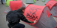 Ein Mann in schwarzer Kleidung klebt ein rotes Poster auf ein graues Zelt, darauf ist ein Herz gemalt, eine Ziffer Zwei und das Wort Block