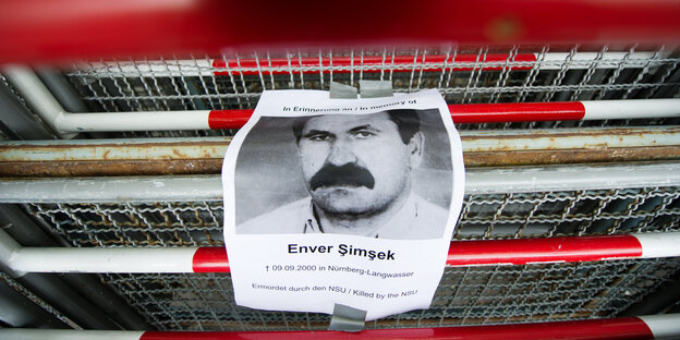 An einem rot-weißen Absperrgitter hängt ein Zettel mit dem Porträt von Enver Şimşek