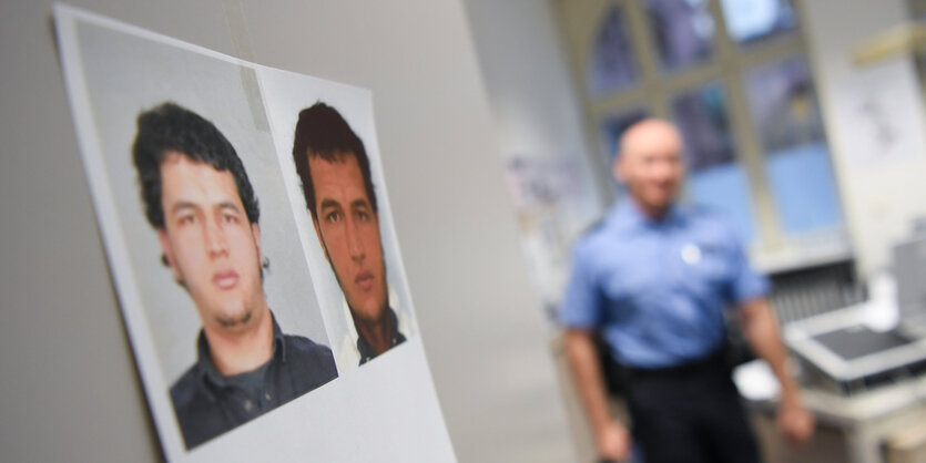 ein Fahndungsfoto von Anis Amri hängt an einer weißen Wand, im Hintergrund ist verschwommen ein Polizist zu sehen