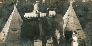 Auf einem lebensgroßen Elefanten aus Pappe sitzen ein Mann und Junge. Eine Dame in feinem weißen Kleid und Hut hält den Elefanten an einer Schnur. Im Hintergrund sind Zelte aufgebaut.