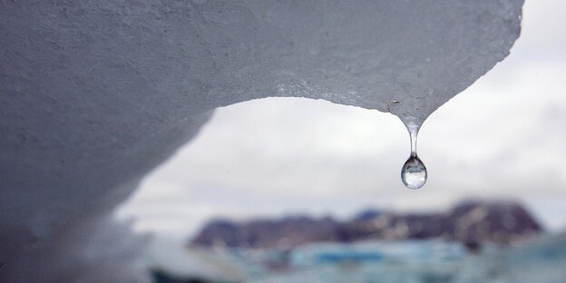 An einem schmelzenden Eisberg hängt ein Tropfen Wasser, im Hintergrund sieht man eisige Landschaften