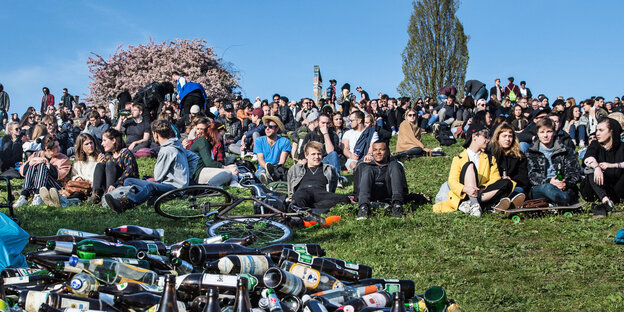 Jugendliche sitzen auf einer grünen Wiese in der Sonne, im Vordergrund stapeln sich leere Flaschen