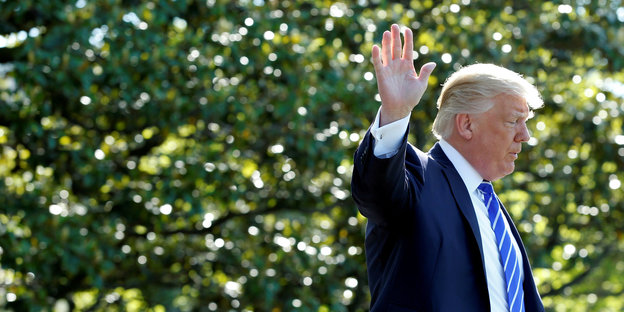 Donald Trump steht vor grünen Bäumen, wendet den Kopf ab und winkt in Richtung Betrachter