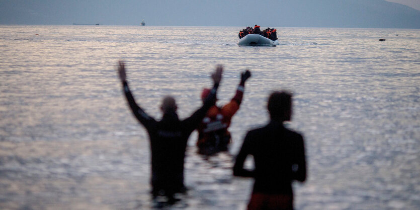 Menschen stehen im Wasser und heben die Arme. Sie warten auf ein ankommendes Schlauchboot.