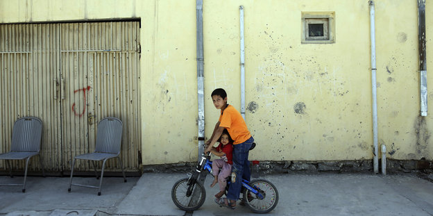 Zwei Kinder fahren auf einem Fahrrad an einem Haus vorbei