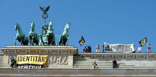 Personen der "Identitären Bewegung" auf dem Brandenburger Tor neben der Quadriga. Sie halten ein Transparent mit der Aufschrift "Grenzen schützen, Leben retten" in die Luft. Ein anderes Transparent ist unter der Quadriga gespannt
