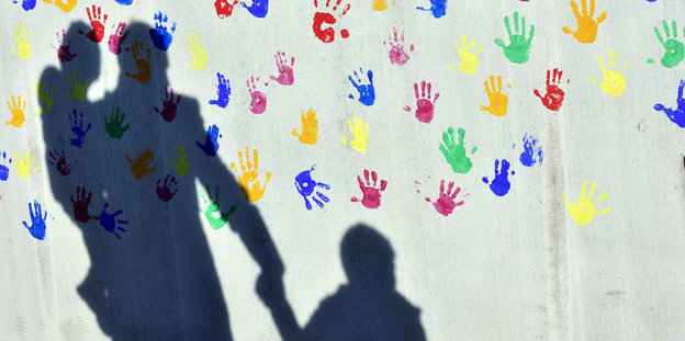 Auf einer Wand, die mit bunten Handabdrücken bemalt ist, sieht man die Schatten von einer erwachsenen Person mit zwei Kindern