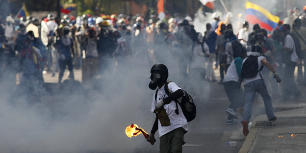 Ein Demonstrant mit Gasmaske vor dem Gesicht hält eine entzündete Flaschenbombe in der Hand. Hinter ihm ist eine Demonstration zu sehen. Rauch verdeckt die Sicht etwas.